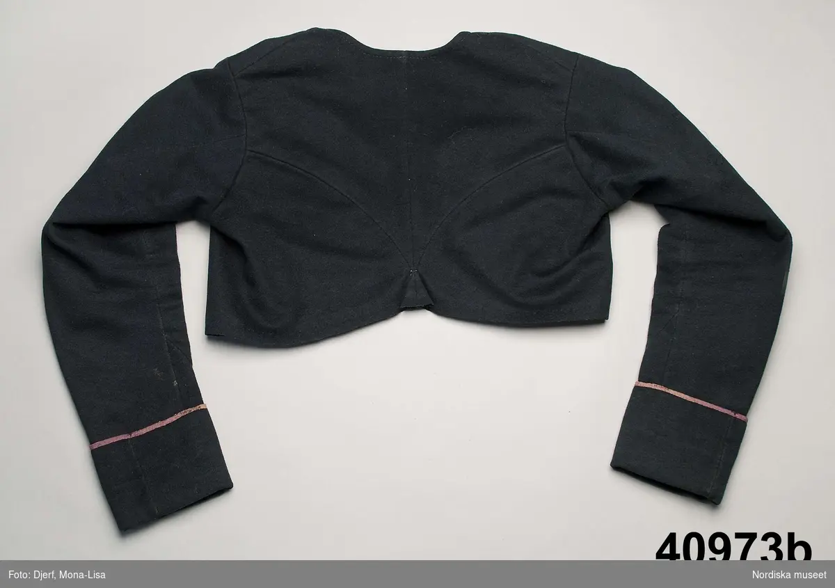 Huvudliggaren: Dräkten "begagnades till början av 1840-talet, linnet av senare årtal."
a.1-2 bindmössa m. stycke
b. tröja längd 35 cm, ärml. 46 cm
c. överdel
d. förkläde
e. kjol
f. 1-2, skor
g. 1-2, bindmössa m. stycke


Kvinnotröja, kort modell av svart kläde,  kallad midjetröja, 2 fram/sidstycken, 2 bakstycken som avsmalnar i ett litet skörtveck mitt bak, nederkanten råklippt och ofållad. framstyckena går omlott med snibb, det högra framstycket har en laskad cirkeldekor mitt fram. Dubbelknäppning med hakar och hyskor fästade genom tyget, isydd lång ärm formad med 2 sömmar, 9,5 cm brett ärmuppslag kantat med lila sidenband. Helfodrad med smalrandig halvbomullskypert med smala ränder på brun botten, de röda och vita ränderna i ullgarn, varpen gråbrunt lingarn, troligen en återbrukad kjol. Tröjan handsydd.
Se Greta Hedlund: Dräkt och kvinnlig slöjd i Ovansjö socken sid 116-119, 1981 där liknande tröjor beskrivs.
/Berit Eldvik 2010-11-09
