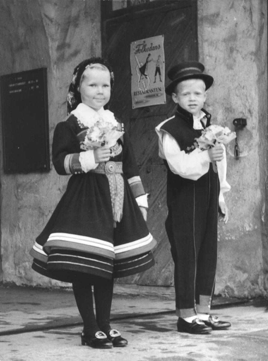 Dronning Fabiola besøker Norsk Folkemuseum i juni 1965. En liten gutt og en liten jente,begge i setesdalsdrakter, holdende hver sin blomsterbukett, venter foran Bergensporten, bygning. nr.301.
