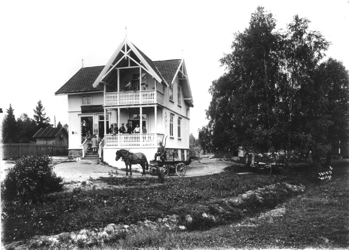 Fredbo landhandleri på Høybråten i Oslo. Eksteriør av huset med folk på verandaen og hestekjøretøy utenfor.