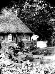 Eldre mann med hatt utenfor gårdsbygning med strå på taket. 