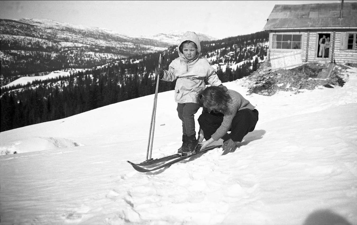Guri og Kari Arentz på Ligardshaugen for første gang påsken 1940. Kari får hjelp av Guri til å spenne på seg skiene. I bakgrunnen ses hytta som er under ombygning.