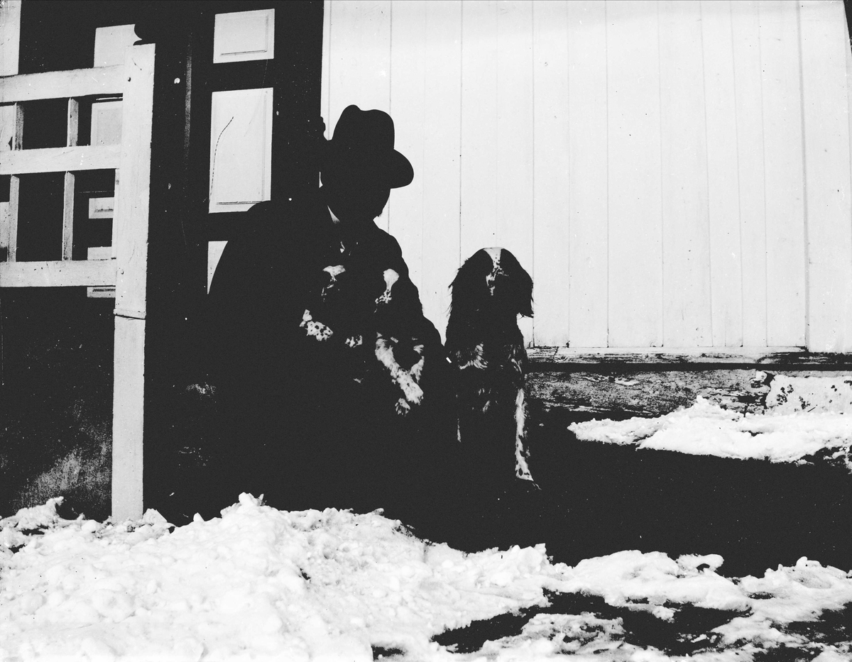 Mann på huk med hundevalper i fanget. Sitter foran en bygning, snø i hagen.