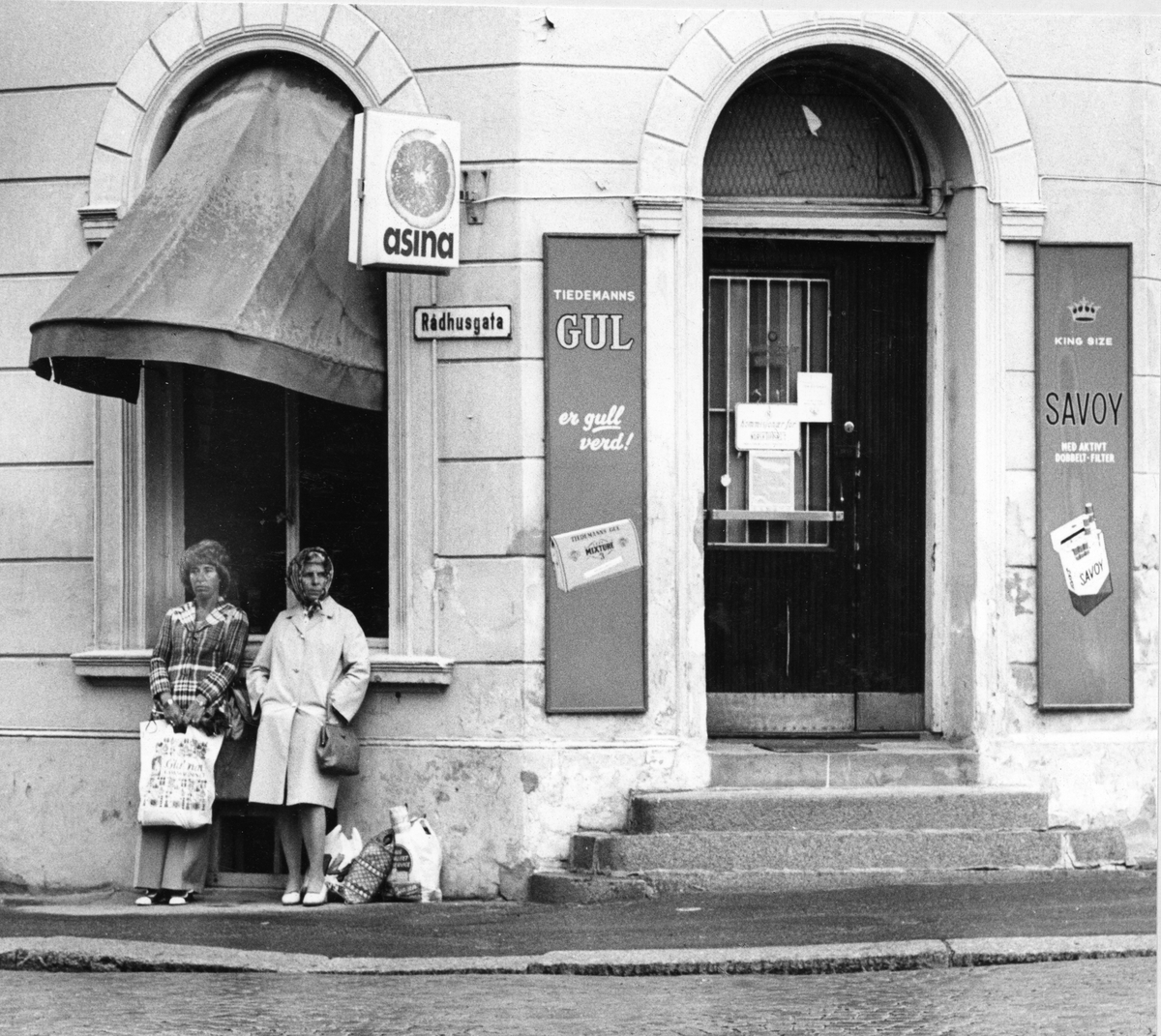 Gatebilde. To damer foran husvegg. Kiosk på hjørnet av en bygård. Reklameskilt for Tiedemanns Gul og Savoy sigaretter.