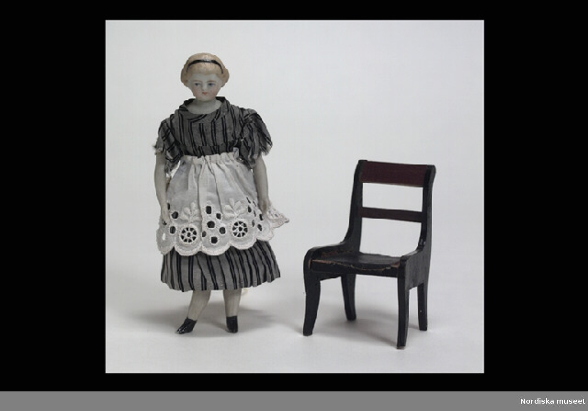 Inventering Sesam 1996-1999:
Docka  H  13    Stol  H  7,5    Barnbädd  L  9,5   B  6 (cm)
Dockskåpsdocka, kvinna, sittande (fastnålad) på stol, med barnbädd och babydocka (a-c). 
a) Docka; huvud, sköld, halvarmar och halvben av biskvi, kropp i stoppat tyg, målade ansiktsdrag och blont hår, svart diadem, grå sidenklänning med svarta ränder, vitt förkläde, underkjol och mamelucker, svartmålade kängor modellerade i massan. 
b) Stol, av svartmålat lövträ, rygg med brunbetsat överstycke och tvärslå, något svängda ben. Under bottnen med blyerts. "Thyra".
c) Stoppad bädd, klädd med vitt bomullstyg, garnerad med spets och rosa sidenband, fastsydd babydocka av biskvi, iklädd rosa dräkt med spetskrage.
Tillhör dockskåp från omkr 1840, inv nr 145.505, inrett 1872 av leksakshandlare W A Bandel, Västerlånggatan, Stockholm, för dottern Thyra, vilken vid överlämnandet till museet själv gjort uppställningen av föremålen i dockskåpet. 
Anna Womack dec 1997