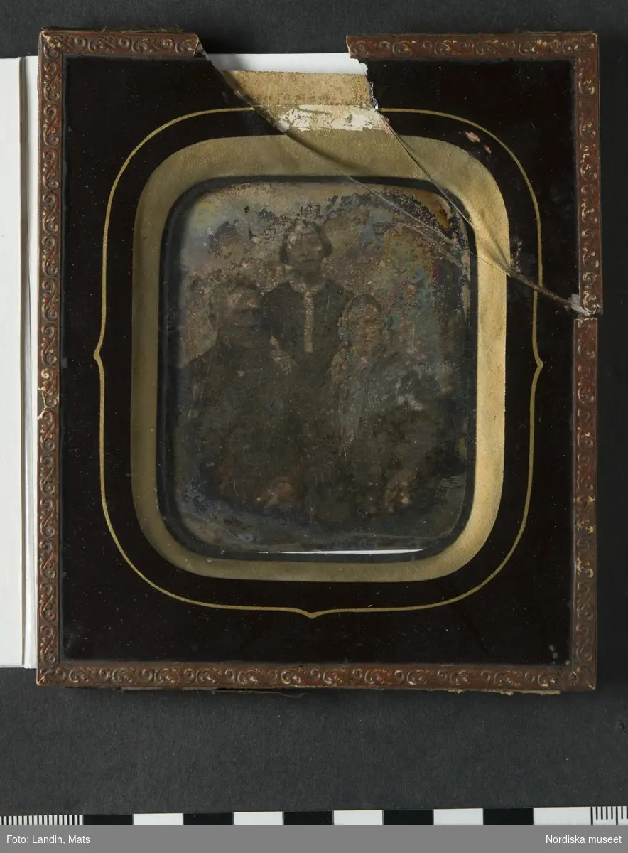 Grupporträtt av 3 personer, föreställer till höger, sittande, brandmästare Gustaf Wilhelm Draminsky, Landskrona och bredvid honom, till vänster, hans hustru Emilia Draminsky (född Hägg), längst bak, stående i mitten, deras fosterdotter, Emilia Fredrika Sophie (1828–1920). Dagerrotyp / daguerreotyp i ram. Plåten oxiderad, glaset sprucket, ramen i lösa delar. Baksida av mörkblå papp med små fastklistrade papperslappar med påskrift: "Mormor/Morfar/tant Emma". Nordiska museet inv.nr 198609
-
Portrait of Gustaf Wilhelm Draminsky, his wife Emilia and their foster daughter Emilia Fredrika Sophie. Oxidized sixth-plate daguerreotype in broken frame.