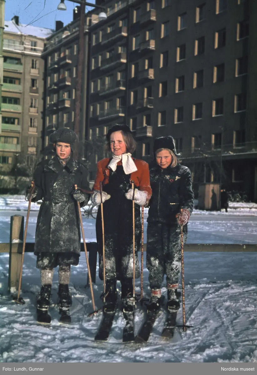 Tre skidåkande flickor framför flervåningshus.
