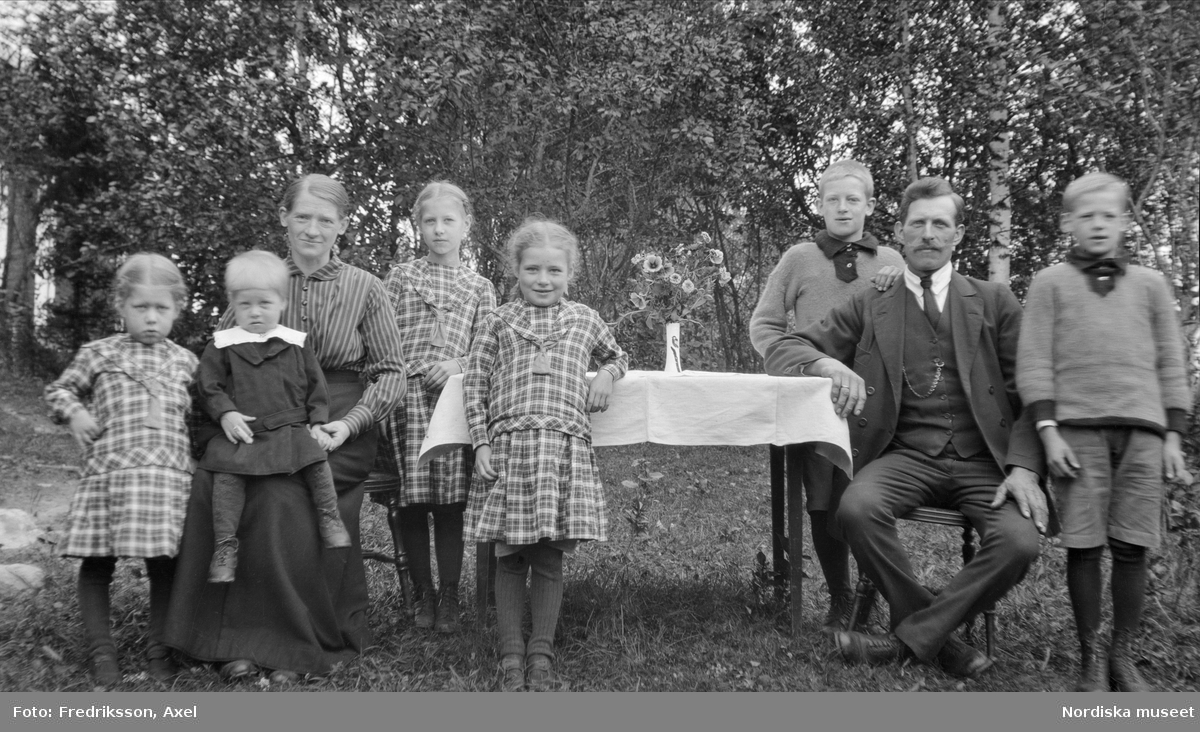Familjeporträtt ute i trädgård. Man, kvinna och sex barn.