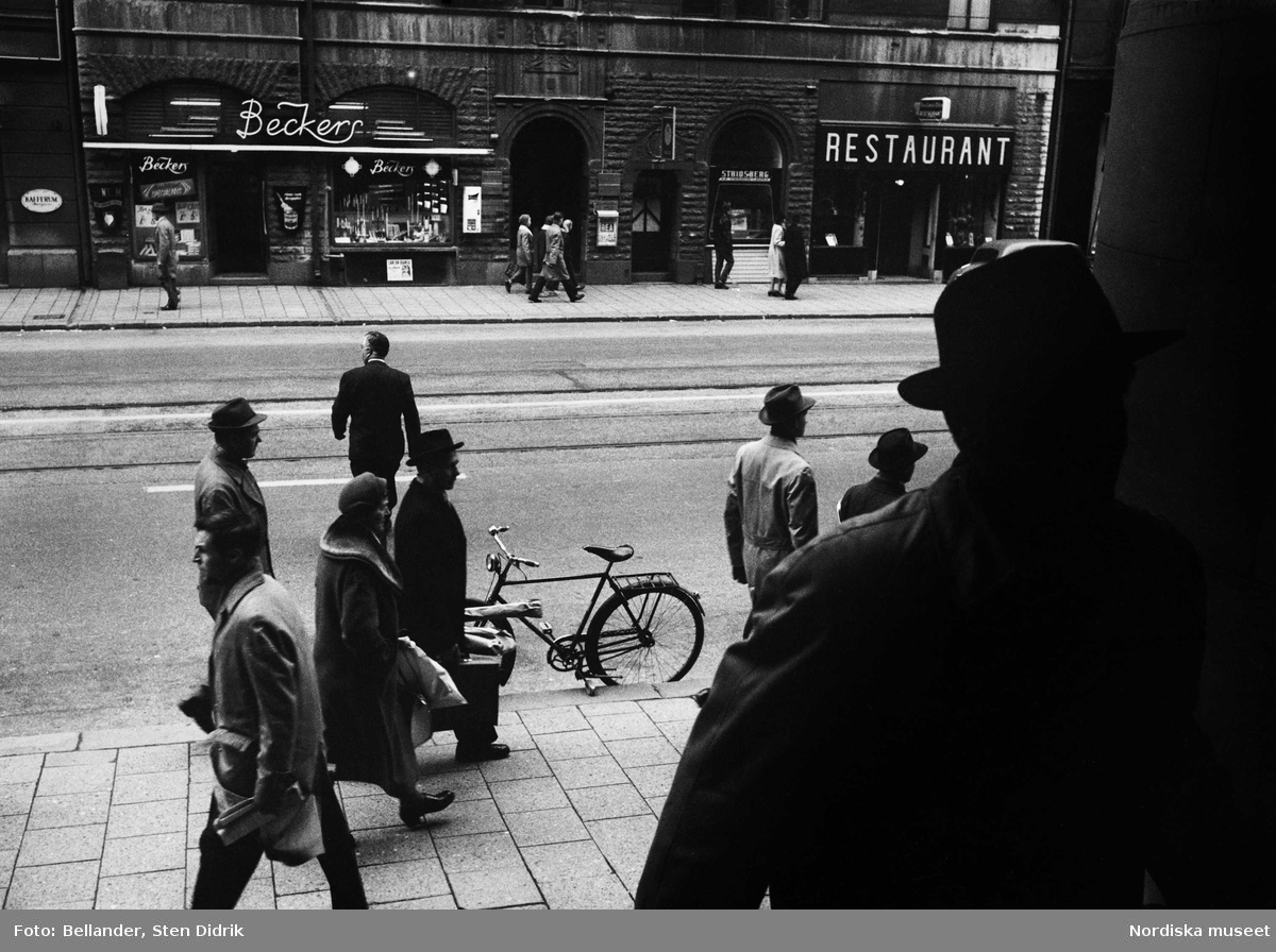 Passerande människor på Kungsgatan.
Män med hatt och rock. En budcykel står vid trottoarkanten.