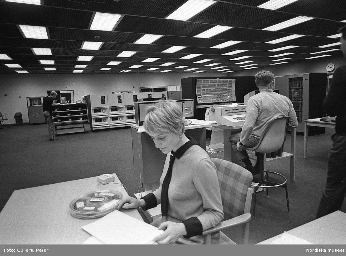 Stockholms Datamaskincentral. I förgrunden kvinna vid skrivbord med magnetband och datormus, man framför tangentbord. I bakgrunden stordatorer.