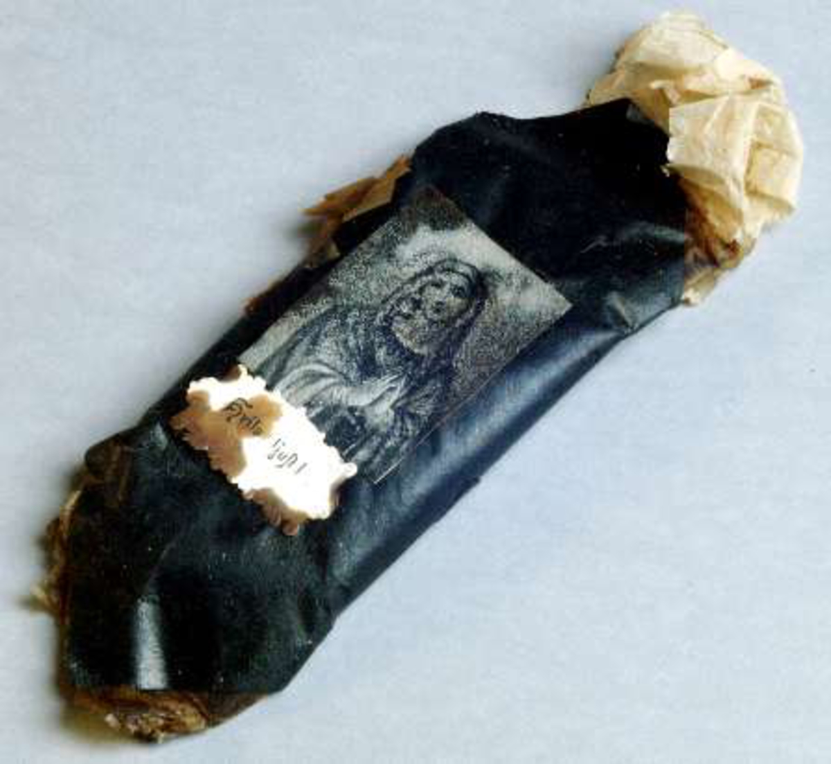 Begravningskaramell med omslag av beige silkespapper och svart glanspapper. Två pappersetiketter, den ena med tryckt bild av jungfru Maria, den andra med tryckt text: Hvila ljust!
