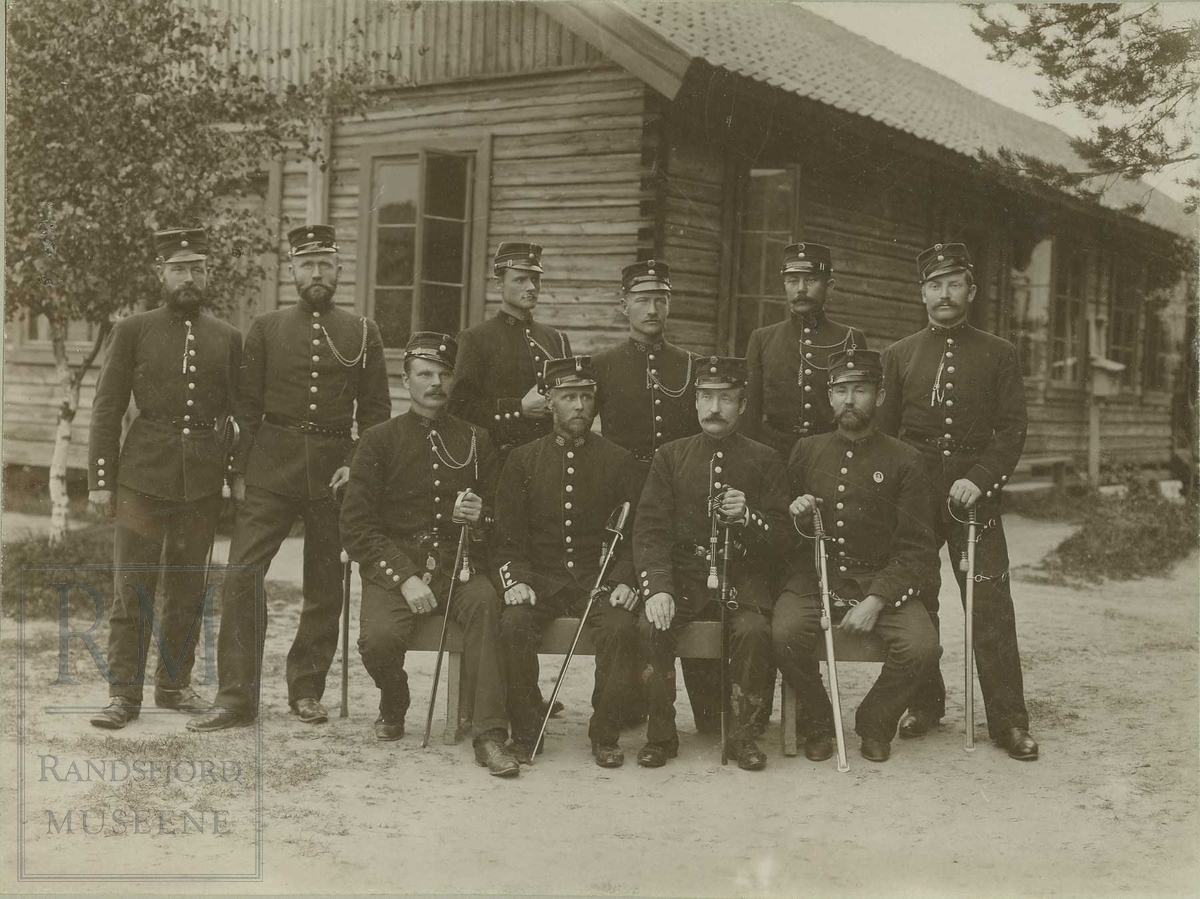 10 soldater med uniform og sabel oppstilt i en gruppe foran ei laftet brakke.