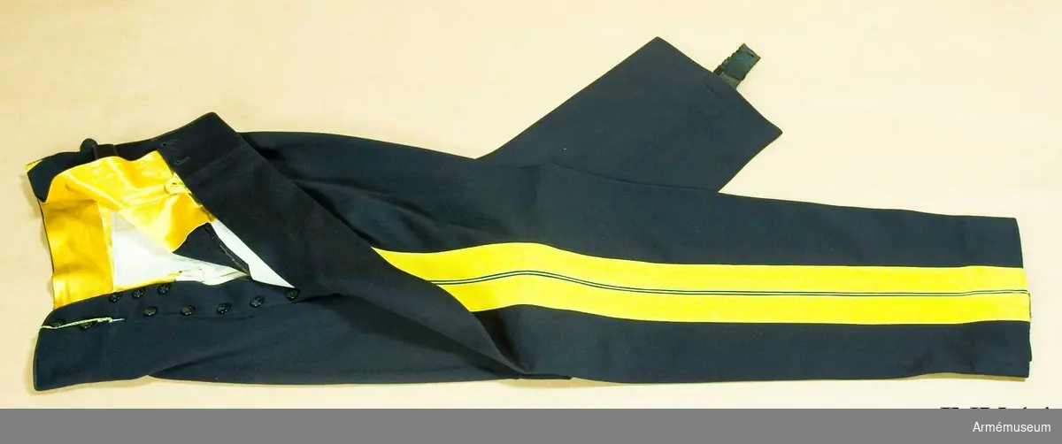 Av mörkblått kläde med dubbla gula klädeslister och  emellan dem en gul passpoil. I gylfen 2 knapprader med 7 resp. 3 svarta knappar av bakelit. Svarta hällor av bomullsresår att knäppas under foten. Gult sidenfoder i linningen.
Har tillhört Gustav VI Adolf (1882-1973).
