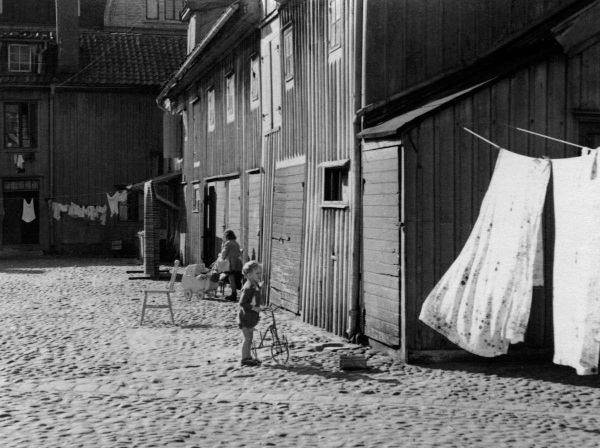 En bakgård med kullersten i Jönköping. Tvätt hänger på tork och två små barn leker med en trehjulig cykel och en dockvagn.