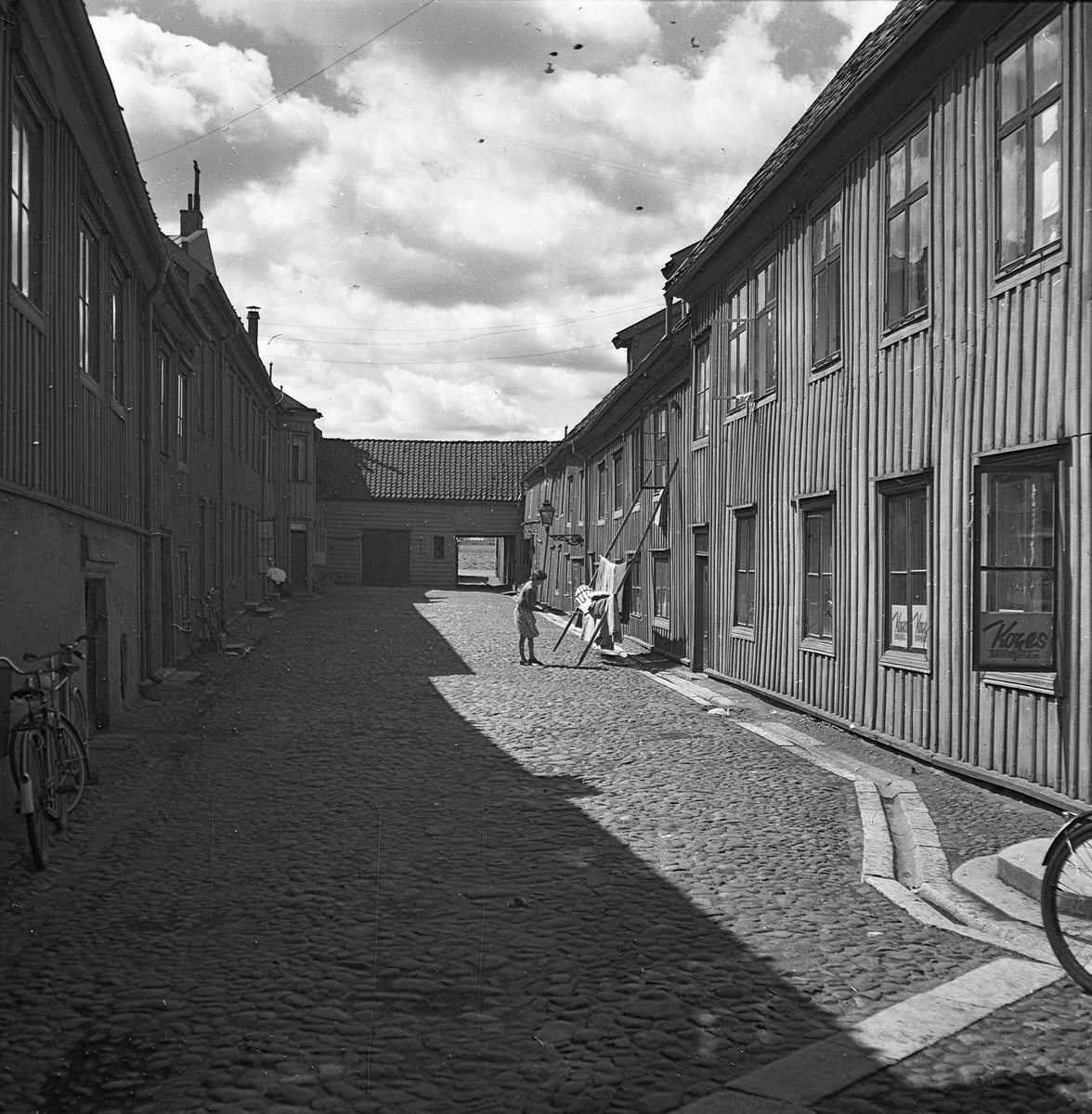 En kullerstensbelagd bakgård i Jönköping. På en ställning lutad mot huset hänger en kvinna upp sängkläder för vädring. I fönstren närmast till höger finns Koges dekorationsateljé som låg här år 1941-1955