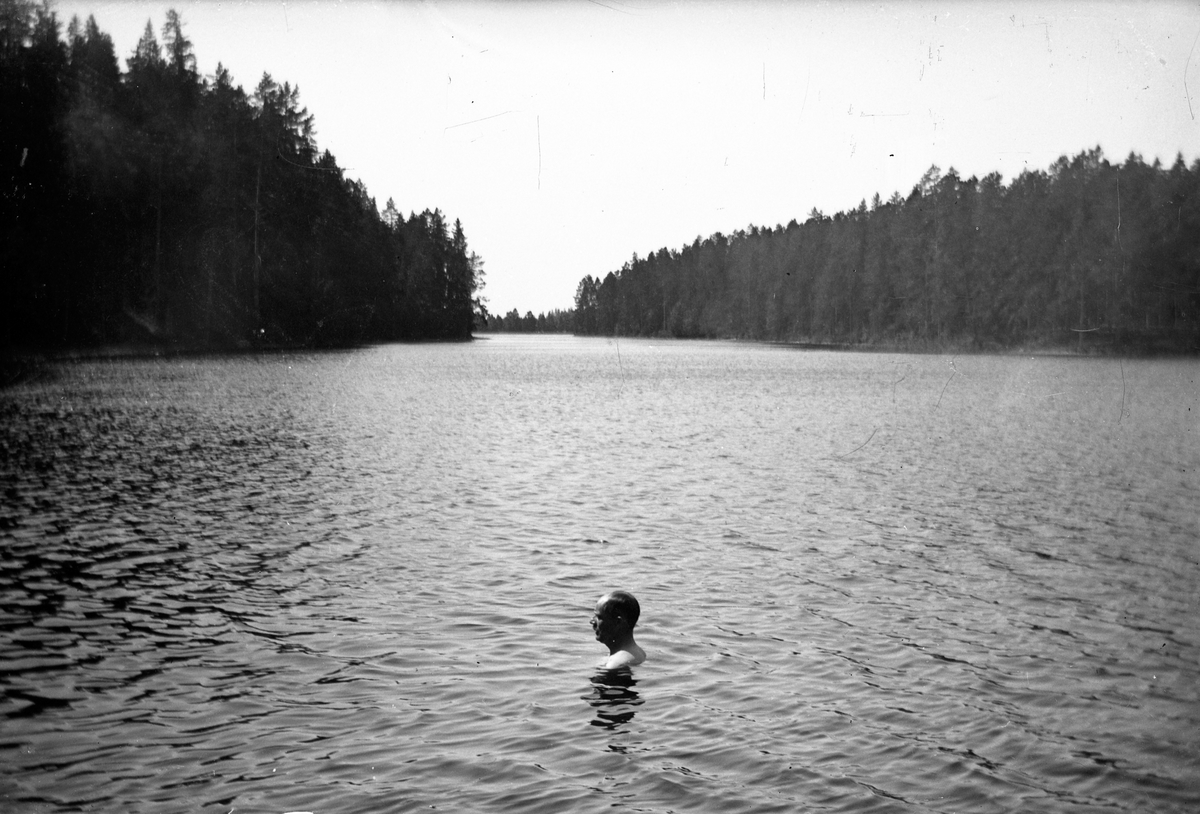 En man med huvudet ovanför vattenytan simmar i sjön vid Lungkliniken i Eksjö. I bakgrunden barrskog.
Fotografens anmärkning: "Mattson i Badet".