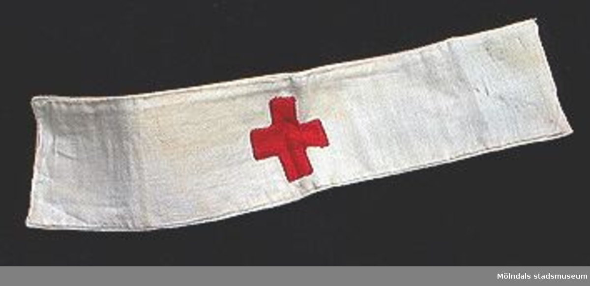 Vit armbindel av två ihopsydda vita tygremsor med ett påsytt rött kors.