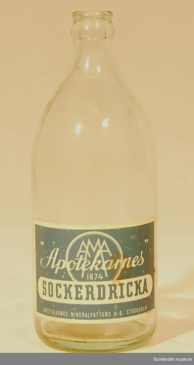 Flaska i ofärgat glas försedd med blå och vit etikett med texten 'AMA 1874, Apotekarnes SOCKERDRICKA, APOTEKARNES MINERALVATTENS A.-B. STOCKHOLM'. Etiketten är tillverkad hos Jönköpings Lit. AB.