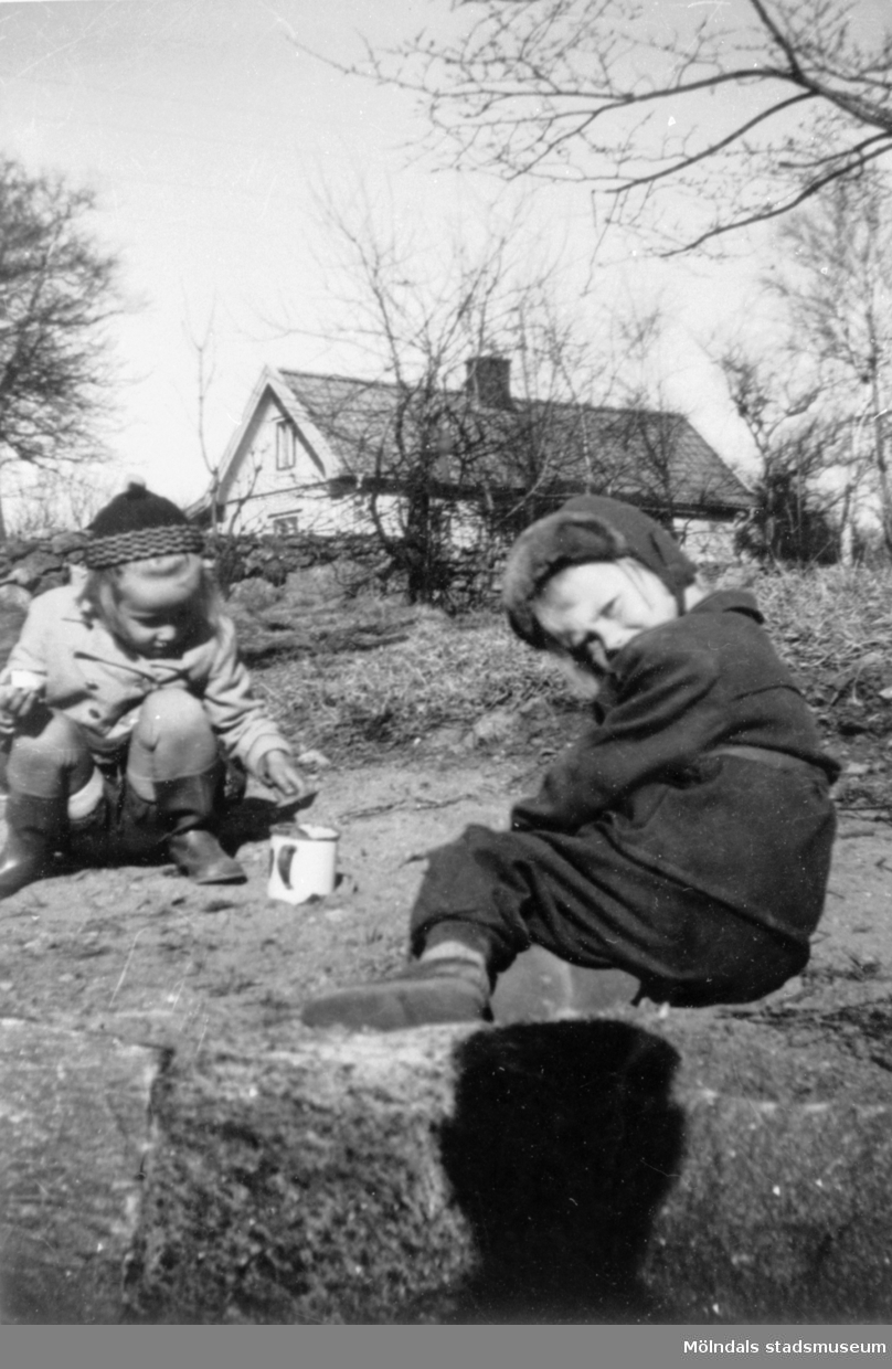 Marie-Louise Lindholm (gift Carlsson) och Lisbeth leker med spade och mugg i trädgården. Flickorna leker på Görjekullen (längst upp på nuvarande Åldermansgatan). I bakgrunden ses Roten K 18, 1950-tal.