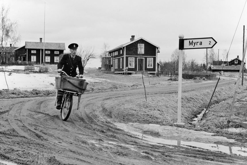 Lantbrevbärare, postiljon Erik Bergman (cykelåkande) i
Delsbo.  April 1956.

Brevbäringsturen är 2 ½ mil lång och omfattar nästan 300 hushåll. Postväskan väger ofta 30-40 kg.
