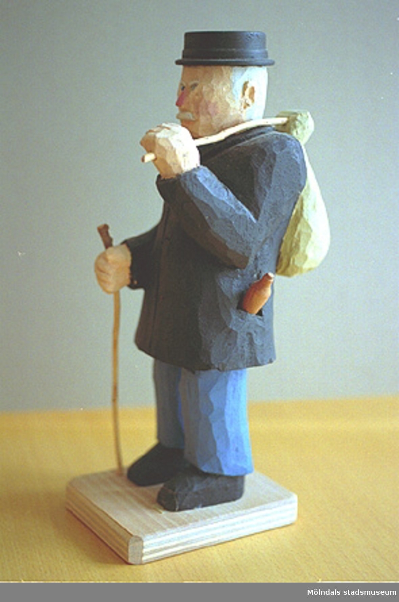 En hattförsedd man som har en påse på ryggen. Han håller en käpp i ena handen. I fickan skymtar man en ölflaska. Harry Bergmans "gubbar" (träfigurer).