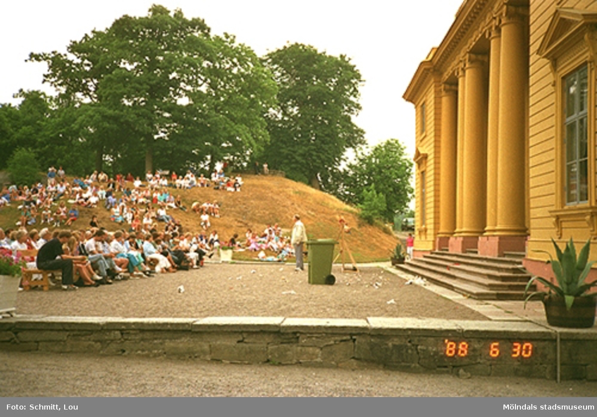 Människor som sitter och tittar på en teaterföreställning framför Gunnebo slottsentré. Till höger ser man en del av det gula slottet.