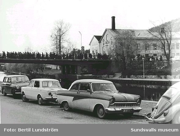 Bilar parkerade på Ågatan, människor på väg över Storbron mot söder. I bakgrunden affischeras en konstutställning arrangerad av Sundsvallskonstnärer.
