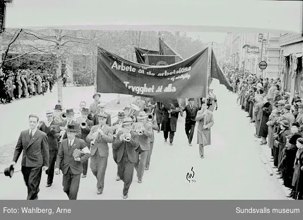 1:a maj-demonstration på väg nedför Köpmangatan mot mötesplatsen på östra Hamnplan, 1927. Fotografiets banderolltexter, "Arbete åt de arbetslösa - ej nödhjälp", "Trygghet åt alla", anspelar på flera förhållanden. Till följd bl.a. av rationaliseringar inom produktionen så var många arbetare inom Norrlands skogsindustrier arbetslösa. Den statliga Arbetslöshetskommissionens dåligt betalda s.k. AK-arbeten ansågs bara fungera som en nödhjälp - man krävde deras avskaffande mot en större samhällelig insats mot arbetslösheten.