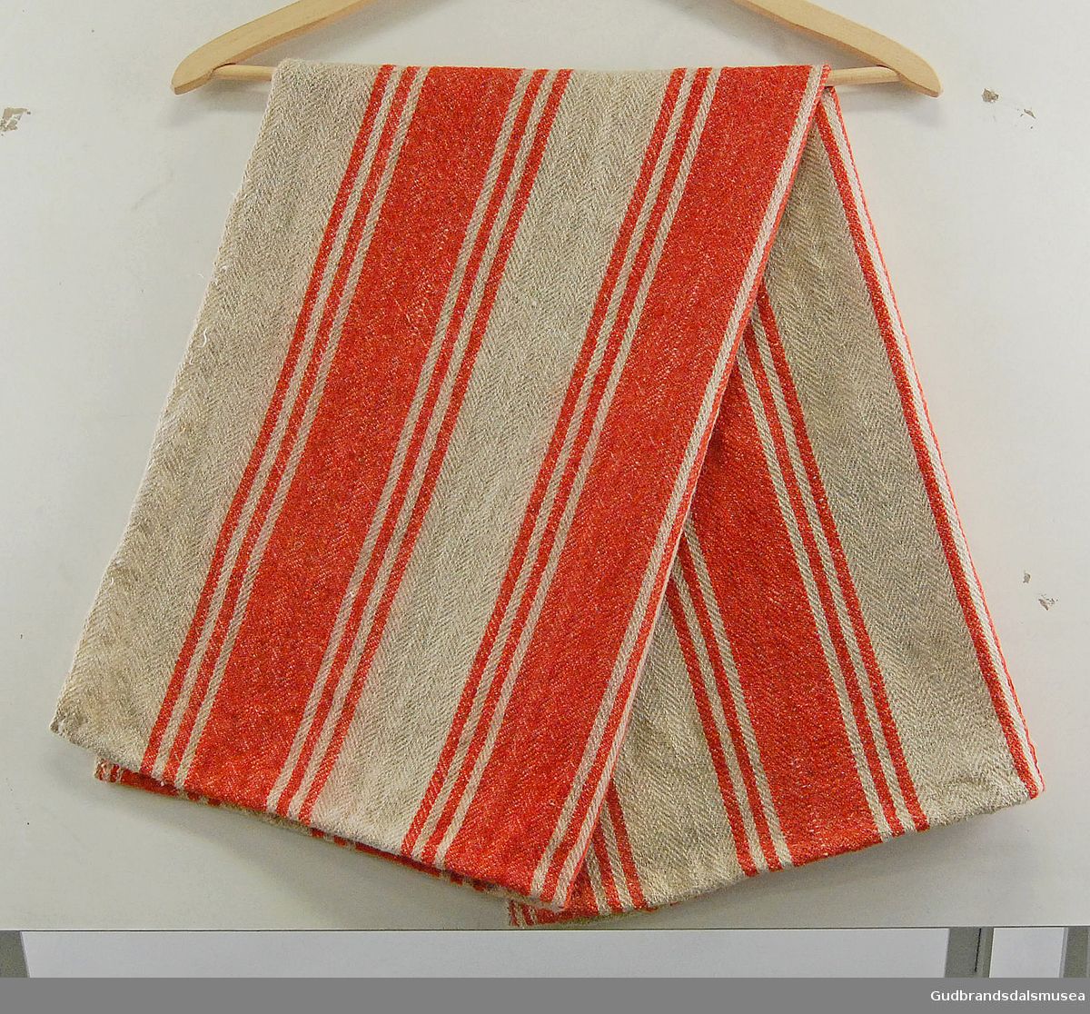 Rektangulært stykke vevd ull med striper i rødt på beige bakgrunn. Innlagt og sydd i kortendene. 