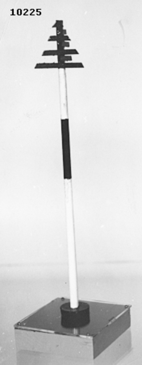 Modell av konprick. Består av en rund stång, målad i två vita och två svarta fält. I toppen en kon, svart. Stången är monterad på en träplatta, botten och sidorna är ljusgrå, översidan blå. Täckt av ett plexiglas.