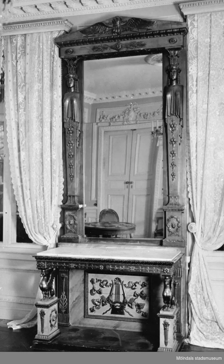 Hög, avlång och dekorerad spegel som står ovanpå ett dekorerat bord. Pjäsen är placerad mellan två fönster. I spegelglaset ses en dubbeldörr med girlangrelief ovanför. Östra förmaket på Gunnebo slott, 1930-tal.