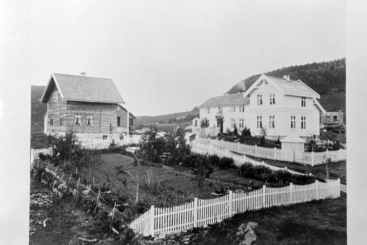 Kaarbøgården fotografert fra veien nedenfor. Til venstre en laftet og umalt stue, til høyre hovedhuset. Hage med hvitt stakittgjerde i forgrunnen.