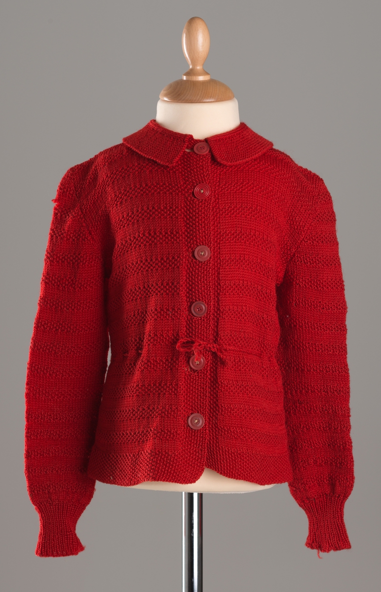 Rød strikket barnejakke i ull. Seks røde knapper i front, fasongstrikket krage, lamge ermer og snøring i livet. Jakka er mønsterstrikket. Se dekorteknikk.