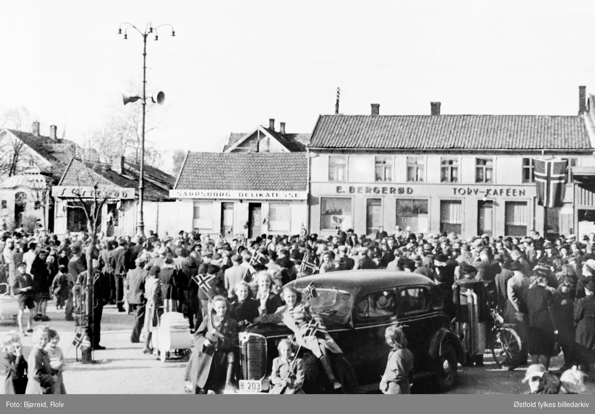 Da freden "brøt løs" i 8.mai 1945.
Fra torvet i Sarpsborg, bil med skilt B-203. Dodge 1938 7-seter med bakmontert vedgassgenerator for knottdrift. Bemerk maiblomst øverst i grillen.Flere forretninger.