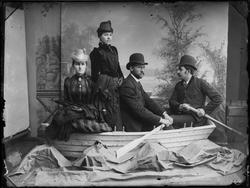 Gruppe arrangert i robåt, inne i fotoatelier. Ingen opplysni