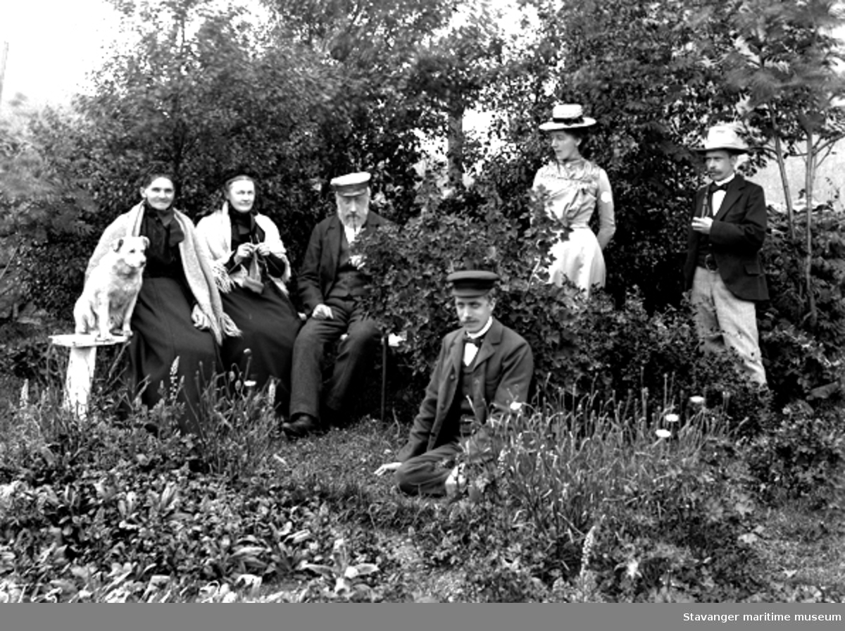 Arrangert foto av mennesker i hage (se ST.S 1981-011-0206), to damer (Julie H. Høier Lund som nr. 2) og en mann sitter på en benk, kvinnen som satt på forrige foto står nå bak en ripsbusk og foran sitter en mann med seilerlue. 