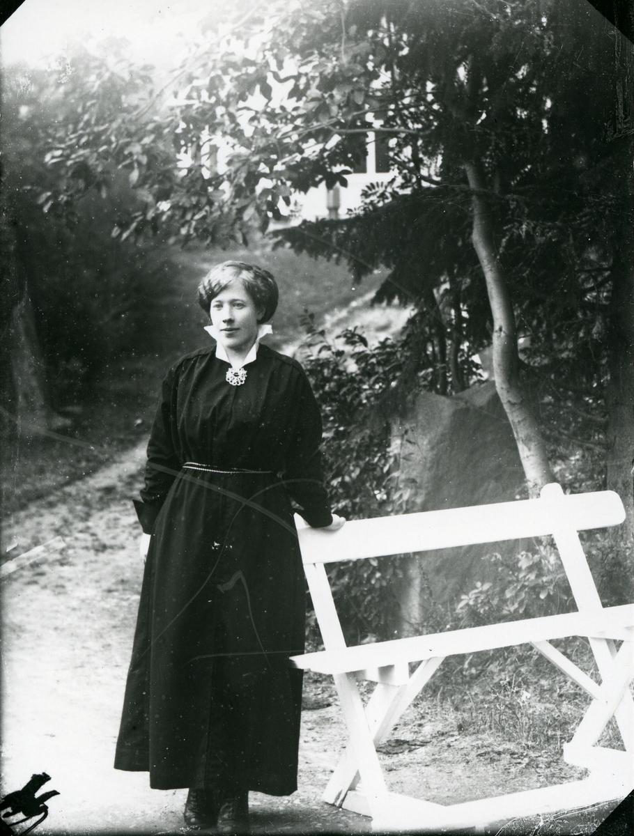 Kvinne ved hvit benk, fotografert på vei med skog og hvit bygning i bakgrunnen