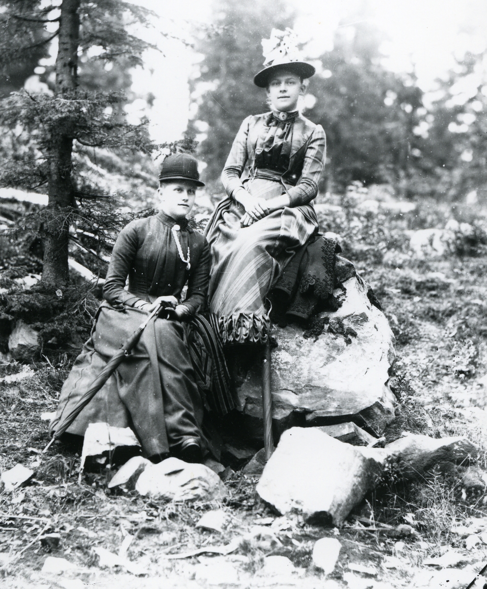 To kvinner i fritidsantrekk, hatter og paraplyer, sittende på steiner i skog