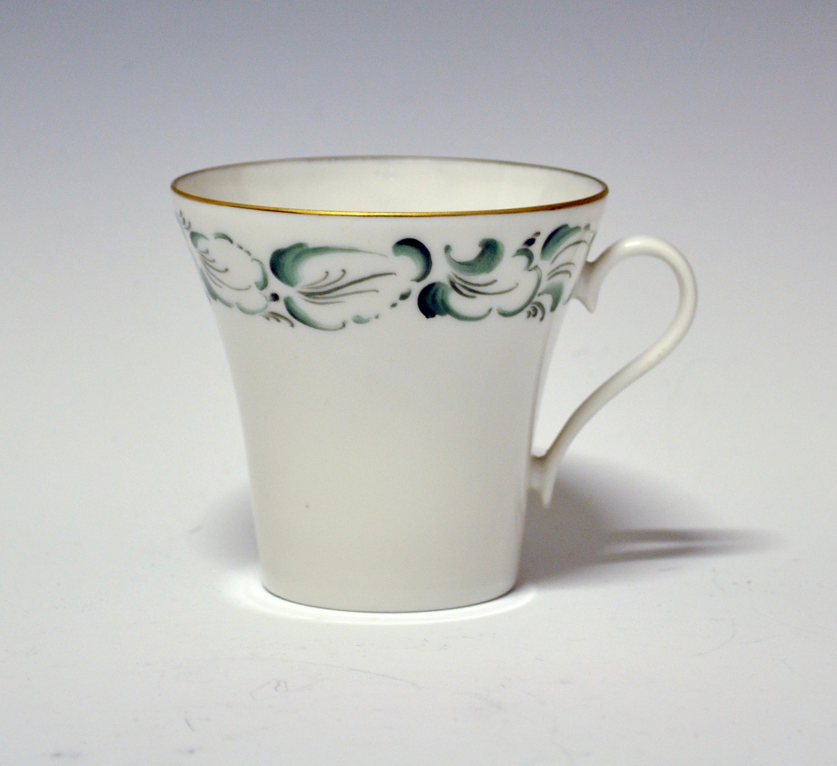 Kaffekopp av porselen. Grønne, stiliserte blader langs kanten. Gullstrek på hank og øverst på koppen.
Håndmalt dekor av Knut Andersen 1975.