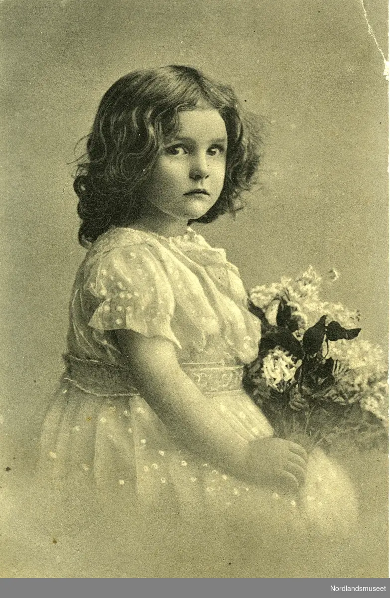 Postkort med portrett av liten jente. Hun har fin kjole med blonder på ermene, og holder en blomsterbukett. Håret er halvlangt og krøllet.

Bakside: Grønt postfrimerke, posthorn, 5 øre. Stemplet Grøtavær, 26.07.1913.