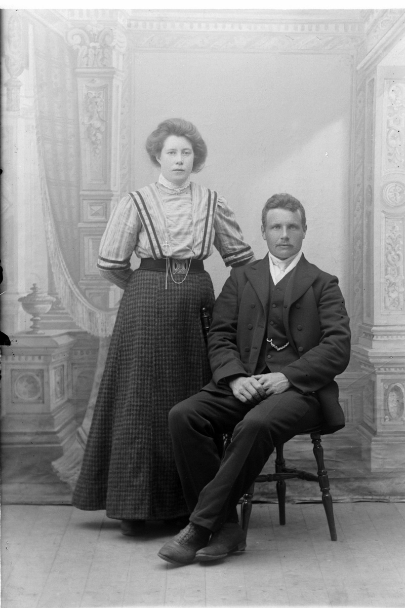 Studioportrett av en kvinne som står ved siden av en mann som sitter.