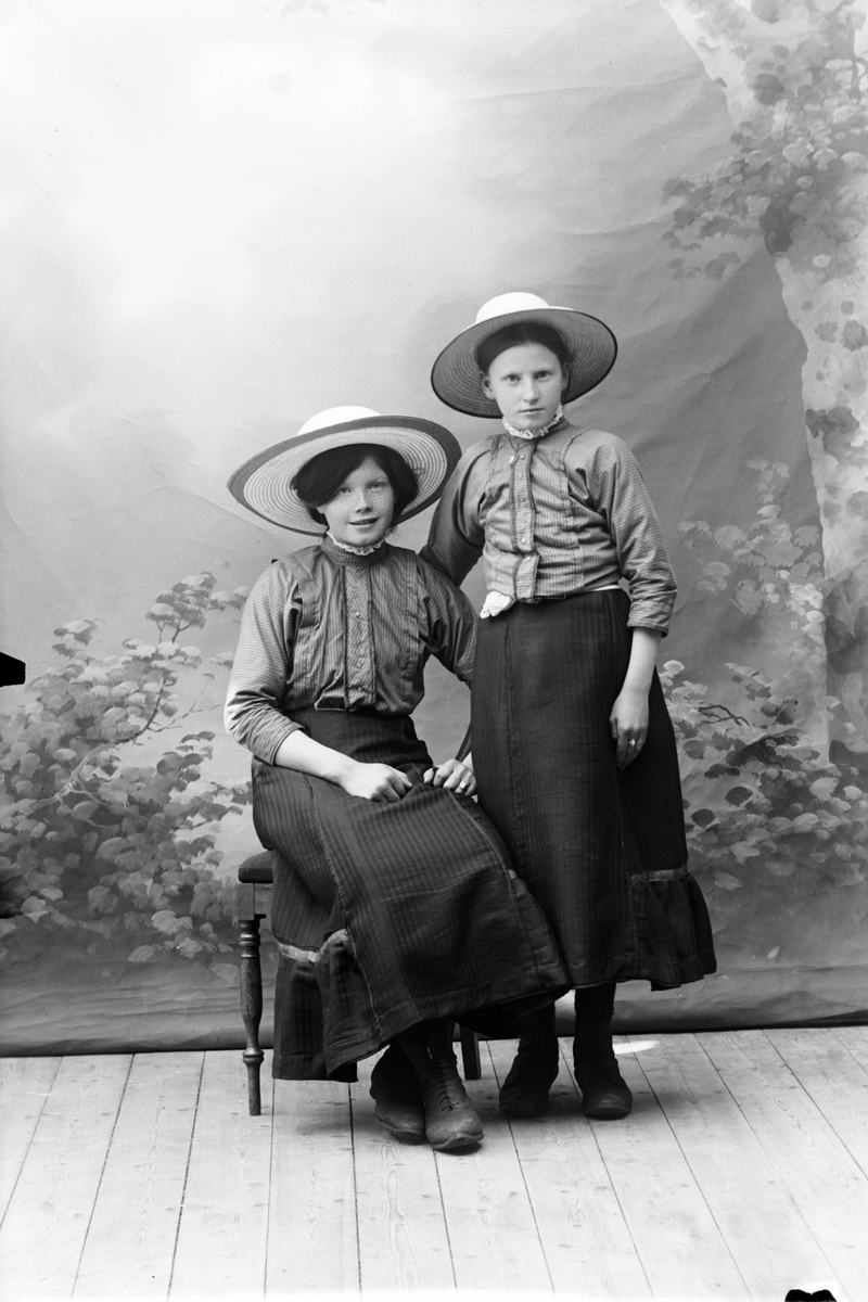 Studioportrett av to unge kvinner med store hatter på hodet.