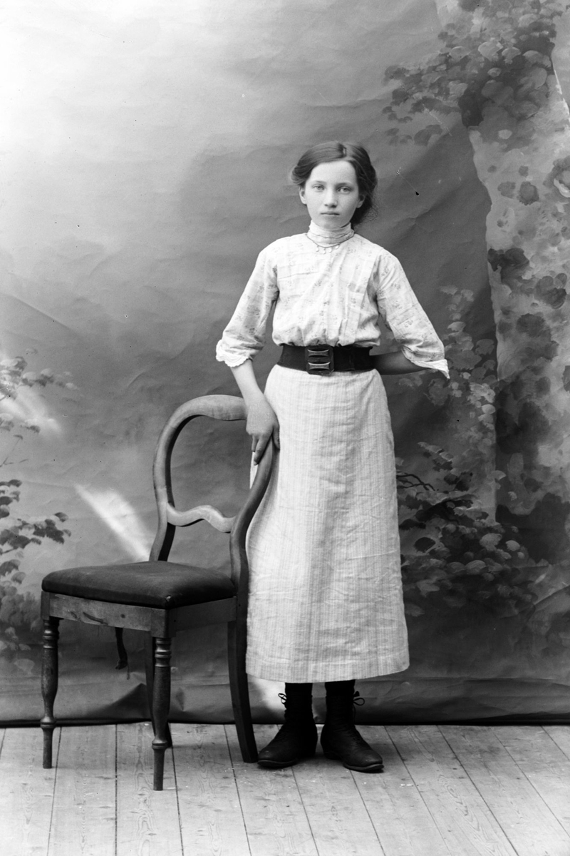 Studioportrett av en kvinne stående ved siden av en stol.
