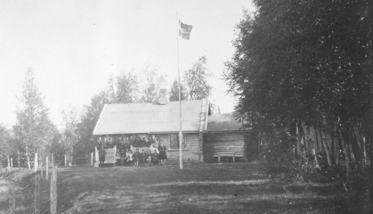 Speidere på Kretsleir i Alta, 1928. I tekst til bildet står det ' Her er hytten hvor speiderne spiste' På verandaen til hytta ses mange mennesker. Flagget er heist i flaggstanga. Det er skog ved hytta. Til venstre i bildet er det et gjerde