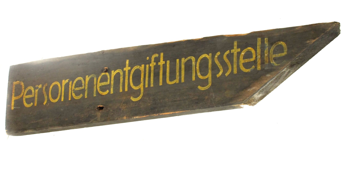 Skilt, av tre, med spiss ende på høyre side, malt i jordgrønn farge med gul tekst på tysk. Noe avbrukket i øvre side.