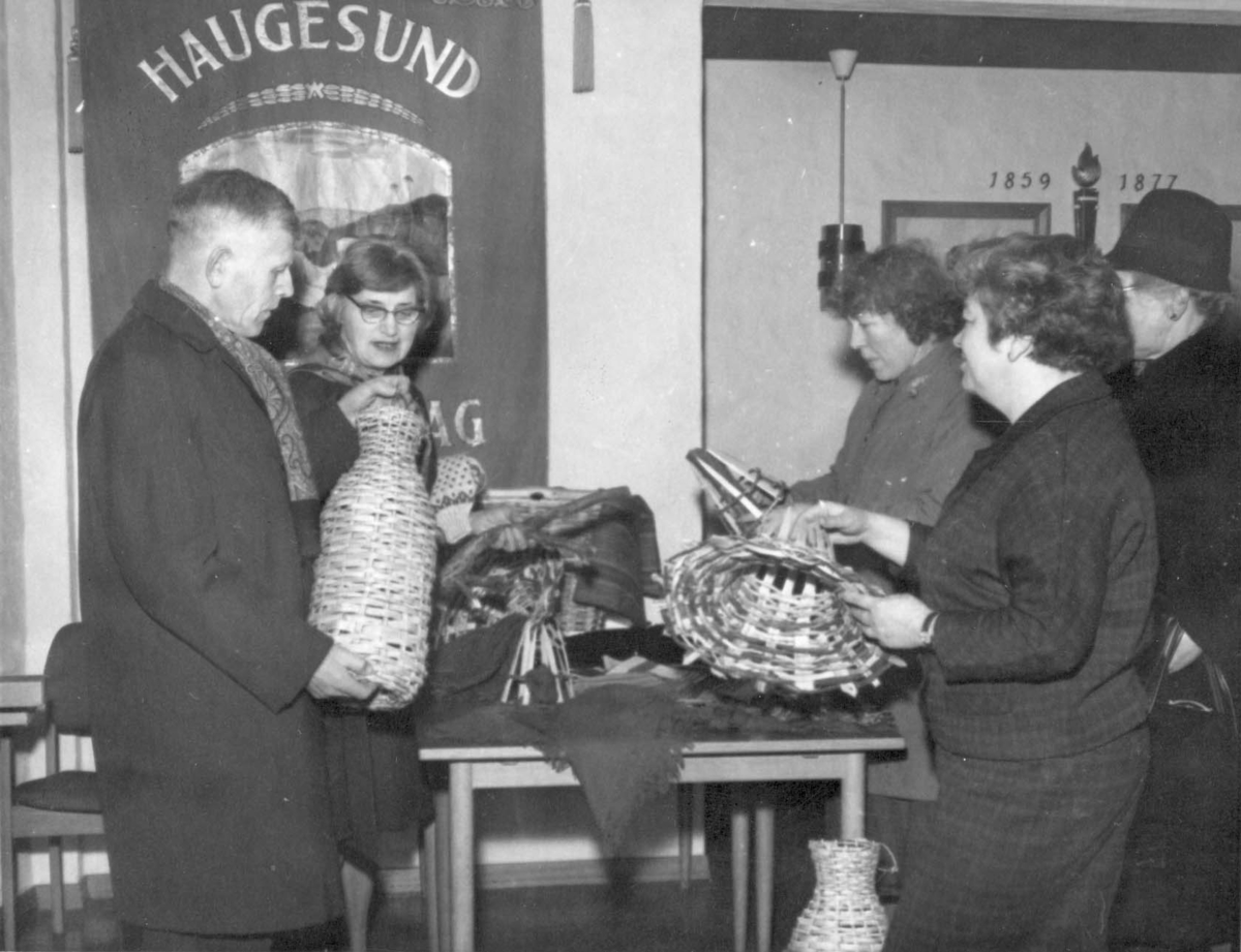 Husflidsdager i Haugesund i DNT-salen. En mann og fire kvinner står rundt et bord og studerer flettede teiner. En fane i bakgrunnen. Åleteiner og "kalvar" til krabbeteiner og "kruner" til hummerteiner, alt kjøpt (utlånt) fra handelsmenn i Haugesund.