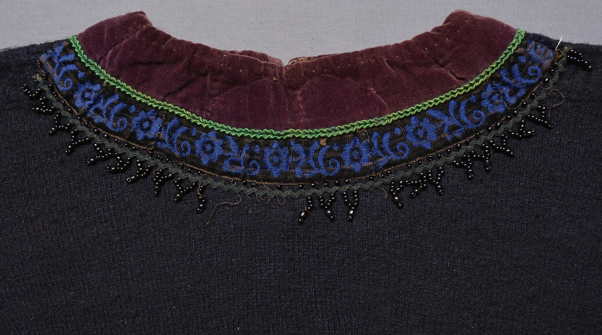 Spedetröja i mörkblått ullgarn garnerad med band, tyg och pärlor i halsringning och vid nederdelen på ärmen. Tröjan är slätstickad med mönsterstickning på axeln och en rätstickad rand i nederkanten. Litet sprund i sidorna. Lila sammetskant (35 mm bred) runt hals och och nederdelen på ärmen, bredvid denna ett smalt grönt garneringsband, sedan ett mönstrat sammetsband i blått på svart botten och ytterst ett svart band med påsydda svarta pärlor.Ärmsprundet hålls ihop av en gulmetallknapp med en infattad grön, facetterad glaspärla, handsydda knapphål.Mitt bak är halsringningen delad av ett 90 mm långt sprund som knäpps med hyska och hake, sprundet är också kantat med lila sammet. Invändigt är halsringning och ärmkant fodrade med halvblekt linne. Foder, band och pärlor är påsydda för hand. Tröjan är valkad.Masktäthet/cm: 4 maskor på höjden och 3,5 maskor på bredden. Axelbredd: 170 mm. Ärmlängd: 420 mm. Bredd nedtill: 340 mm. Märkt invändigt bak vid halssprundet med påsydd tyglapp med texten: "N° 165. a".