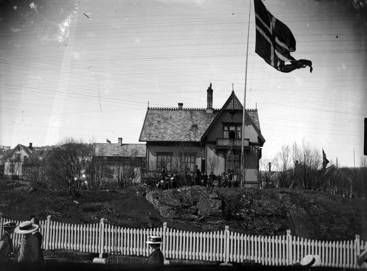 Flagg og soldater (divisjonsmusikken?) foran Generalboligen i Harstad sentrum. I forgrunnen et hvitmalt gjerde.