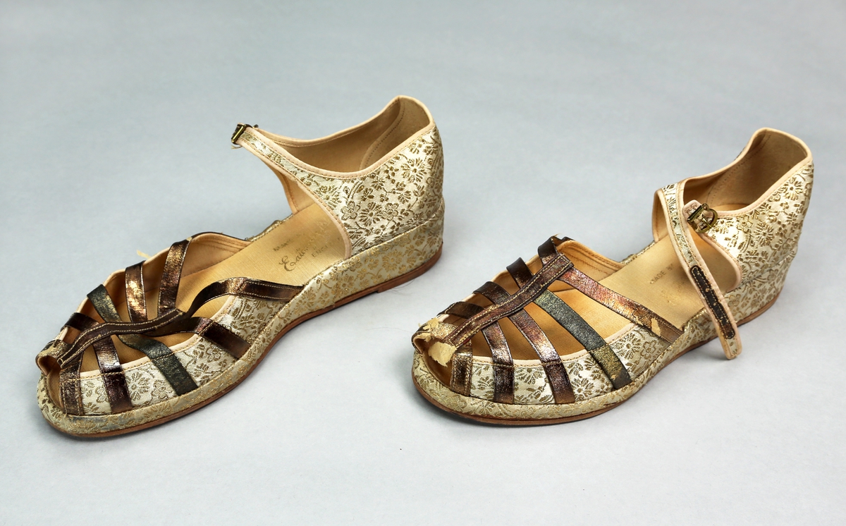 Skopar i eske. Skoene er trukket med stoff som er gullfarget med blomstermotiver i stoffet, og har reimer av bronsefarget stoff over vrista. Sålen er av gummi. 