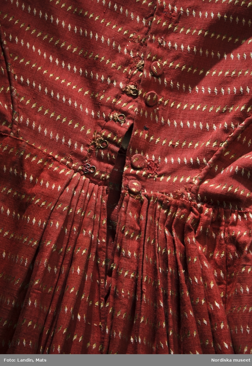 Klänning, kvinnlig dräkt, Stockholm, ca 1820. Bomull, silke, bomullsvadd. 
Klänning i empirmodell av hemvävt bomullstyg med mönsterränder av små sneda rutor i silke i schatterande färger. Det korta livet har applikationer av vaddfyllda "löv" och snedremsor. Ärmen snäv, skuren på snedden med likadana fyllda "löv" vid handleden, och en pålagd puffärm med flikig kant. Kjolen är slät med vidden samlad bak i rynkor. Vadderade snedremsor och större "löv" av avigvänt tyg. Kjolfållen är vadderad till en valk som styvar upp nederkanten. Sydd för Eva Plagemann, f. Åslund (1785-1853), gift 1822 med apotekaren Carl Johan Plagemann som grundat apoteket Nordstjärnan i Stockholm. Familjen är omtalad på flera ställen i Årstafruns dagbok.
