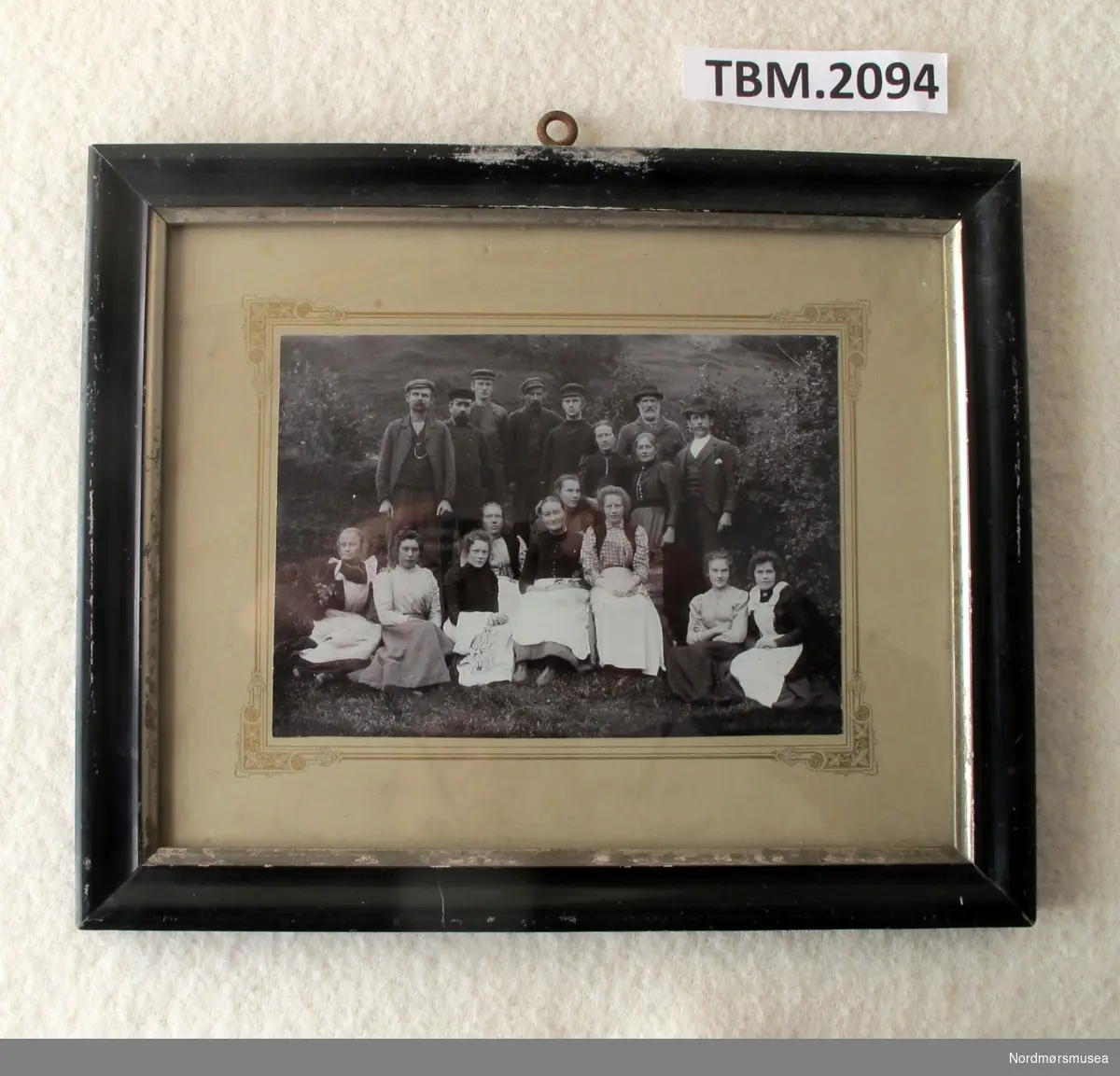 Fotografi av arbeidarane ved Tingvold Uldvarefabrikk oppstilte ute i naturen. Bildet er festa til ei papp-plate med enkel dekorasjon og innrama i ei svart rame.